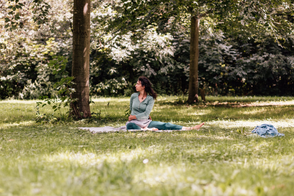 Die Yogalehrerin Maria Lichtenberg verweilt im Sitzen mit gegrätschten Beinen - das eine Bein angewinkelt, das andere ausgestreckt - in einer Drehhaltung in der freien Natur (in der Nähe ihres Yogaraumes in Leipzig).